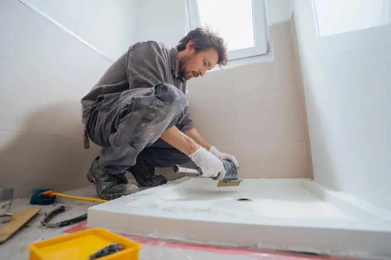 Un homme prépare le sol pour installer un receveur de douche. Il utilise une ponceuse pour lisser la surface, une étape clé pour savoir avec quoi coller un receveur de douche.