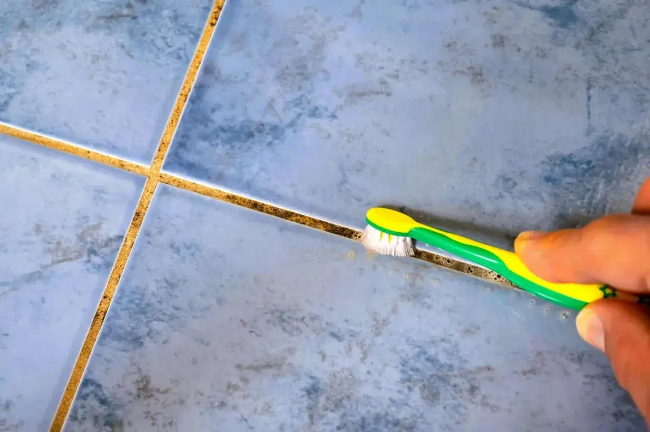 Main tenant, une brosse à dents verte et jaune frotte les joints de carrelage gris pour montrer comment enlever le noir sur les joints de carrelage.