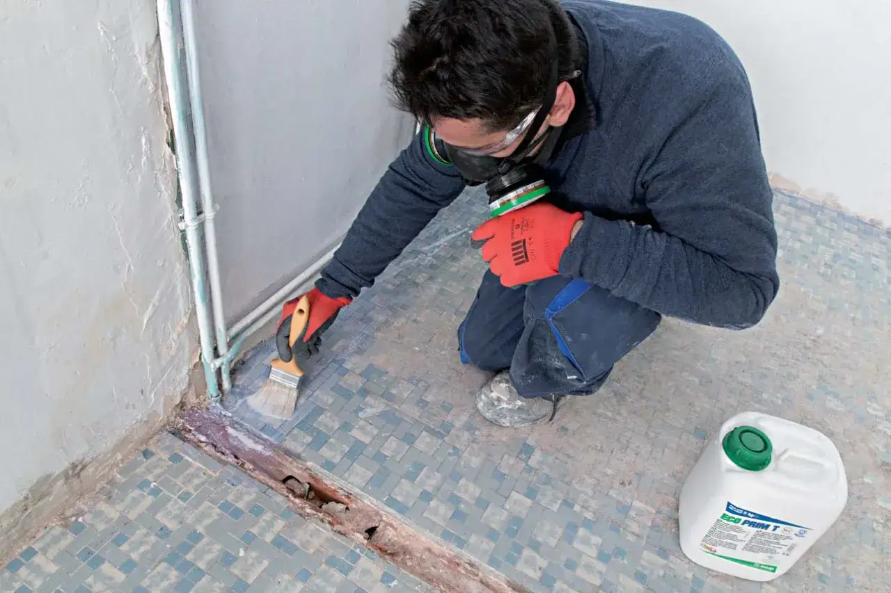 Un ouvrier applique un primaire avant ragréage autonivelant sur un sol en mosaïque usé.