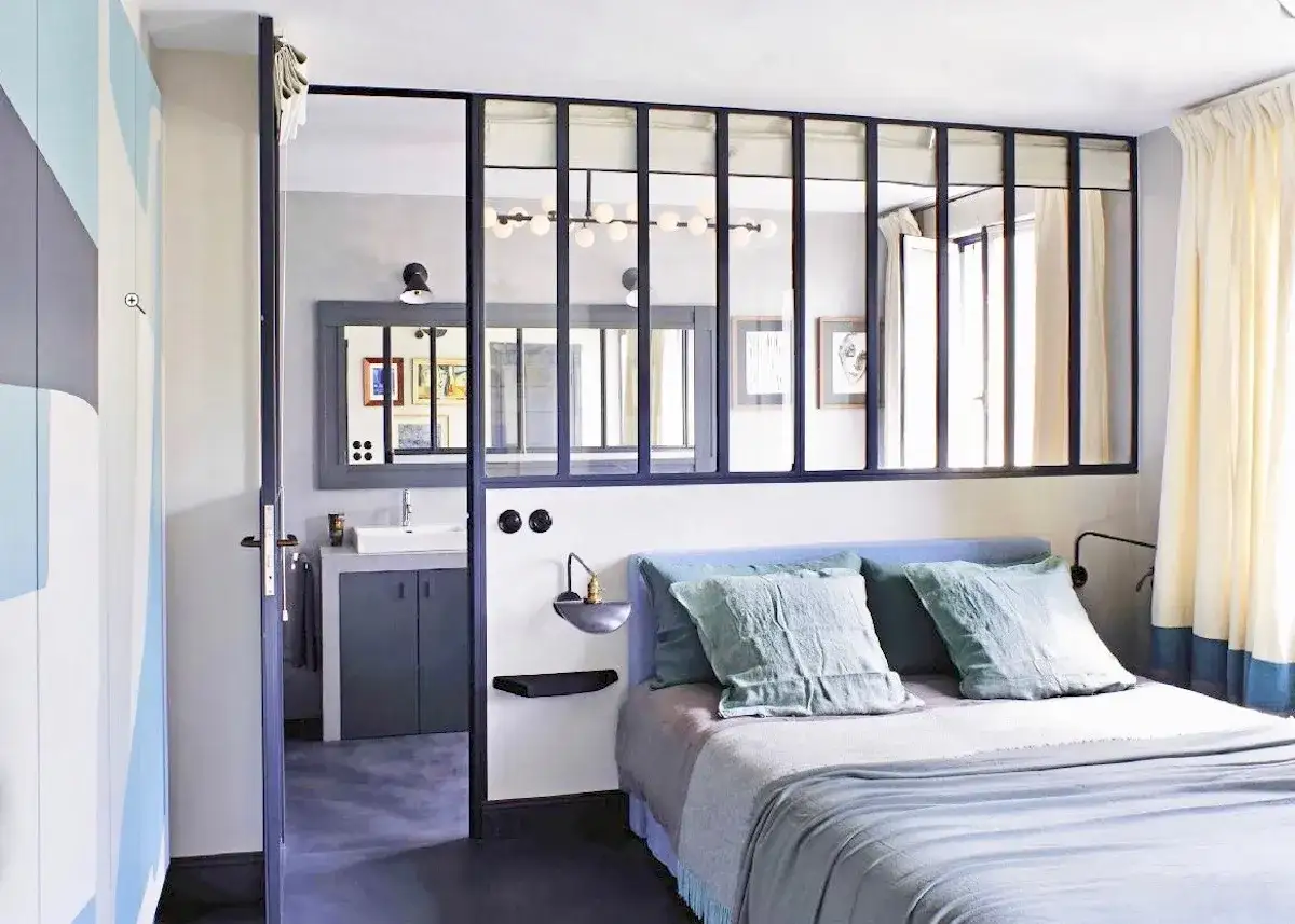 Chambre avec salle de bain ouverte sur chambre séparée par une verrière. Lit bleu, coussins verts, rideaux clairs.