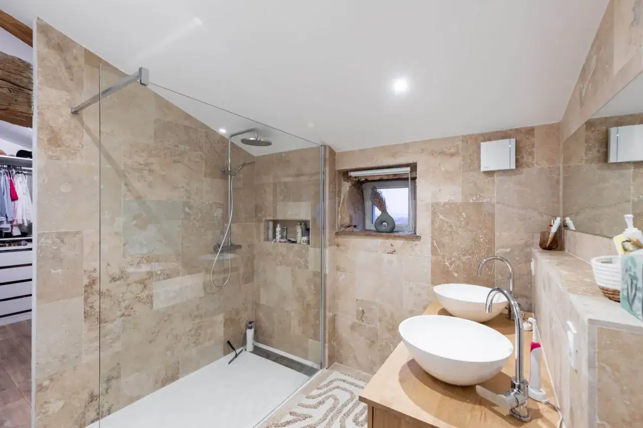 Salle de bain moderne avec douche &agrave; l'italienne, double vasque et carrelage effet travertin. Id&eacute;al pour refaire une salle de bain budget avec style.