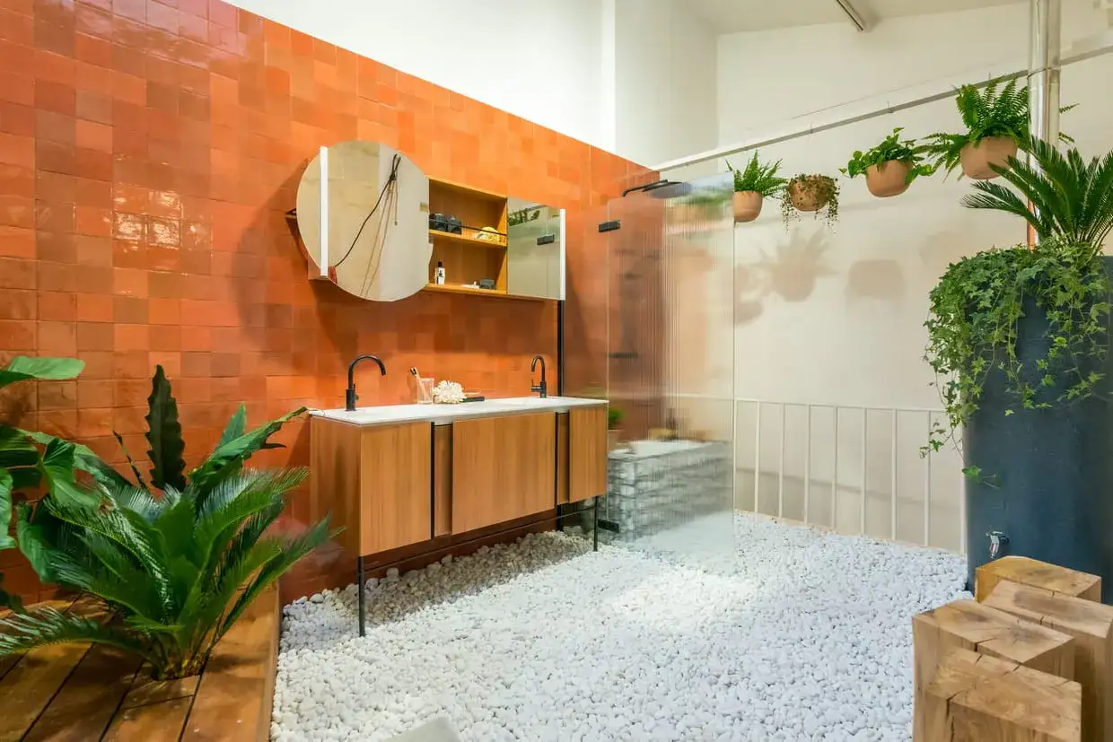 Une salle de bain boh&egrave;me avec murs en terre cuite, double vasque en bois, sol en galets blancs et beaucoup de plantes vertes.