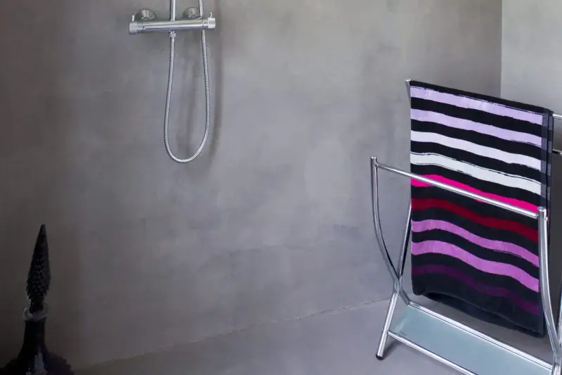 Salle de bain béton ciré aux tons gris, avec une douche chromée et un porte-serviettes orné d'une serviette rayée.