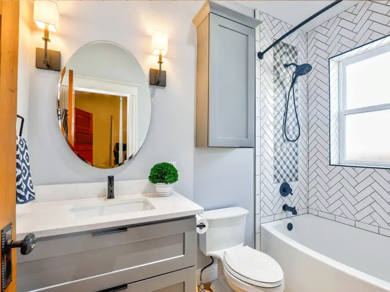 Une petite salle de bain moderne avec baignoire, lavabo gris, miroir rond et carrelage chevrons. Une belle idée pour optimiser l'espace.